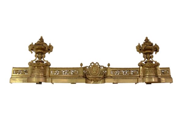 Antique brass fireplace fender with ornate French styling and two decorative pedestal finials, shown straight on.