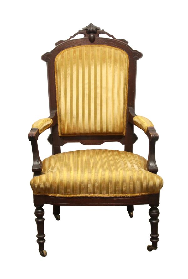 Front view of an Eastlake rotary chair with a dark wood carved frame, yellow striped upholstery, cushioned armrests, and wheels on the front legs.