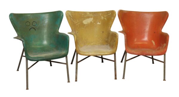 Three fiberglass chairs with steel legs in green, yellow, and orange, shown front view with worn surfaces and a sad face drawn on the green chair.