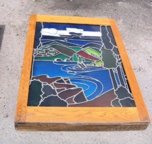 Backlit stained glass window in a dark wood box, showing a multicolor pastoral scene with hills, water, and clouds.
