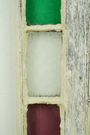 Close-up of an antique Queen Anne stained glass window with three vertical panes in green, frosted clear, and red, framed by worn painted pine, with the glass intact.