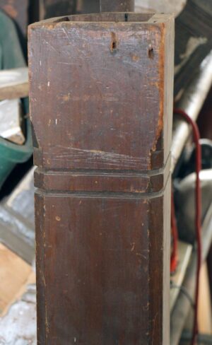 Square wood staircase newel post in a dark wood tone, shown in close-up with a worn top section and visible surface wear.