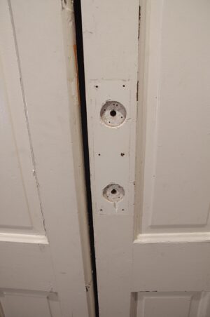 White wooden privacy door with four vertical panels shown in close-up, featuring a narrow center seam and two empty circular hardware openings on the latch side.