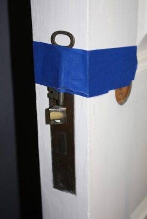 Close-up of a white pine passage door showing the original brass mortise lock and skeleton key, with blue tape wrapped across the latch area.
