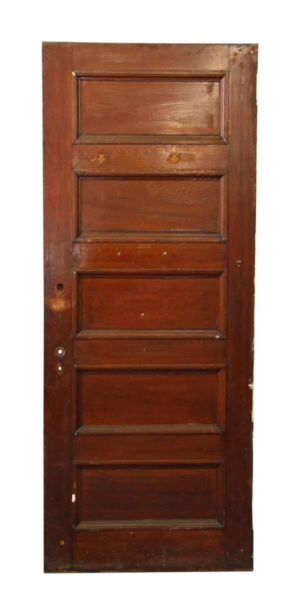 Vintage wooden privacy door with five recessed panels in a dark wood tone, shown full length with visible hardware holes and wear on the surface.