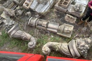 Pair of gray terra cotta cornucopias with carved scroll and fruit details, shown as separated architectural pieces laid out on the ground.