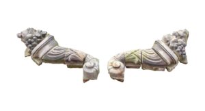 Pair of gray terra cotta cornucopias shown separately, with carved fruit and leaf details and curved segmented forms.
