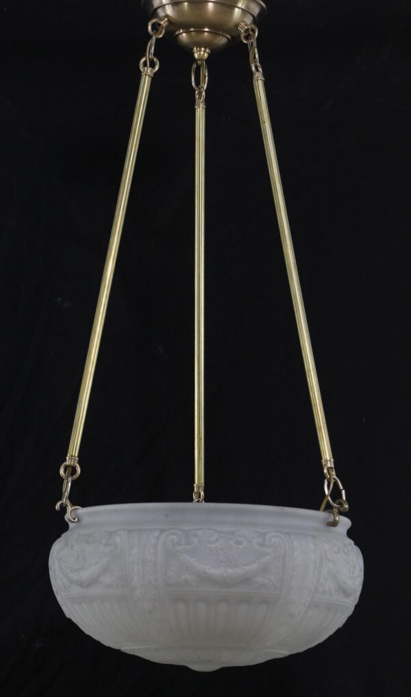White cast glass dish pendant light suspended from three antique brass poles with a brass canopy and ornate relief detail on the bowl.