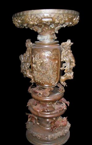 An ornate bronze urn with raised figural dragon decoration and a wide decorative rim, shown in a front view on a plain dark background.