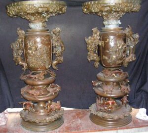 Pair of ornate bronze urns shown side by side, each with raised figural decoration, dragon handles, and a sculpted cover topped by a samurai figure.