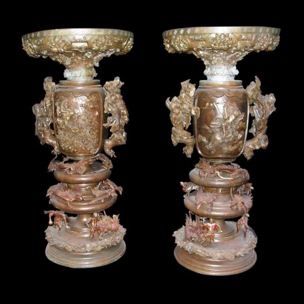 Pair of ornate bronze urns with raised figural designs, dragon handles, and a large pedestal base, shown as a side-by-side pair.