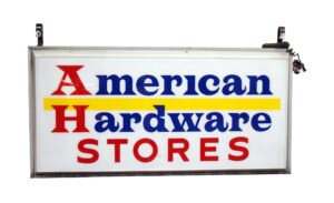 Vintage hanging store sign with the words American Hardware Stores in blue and red on a white panel, with a yellow stripe and hanging brackets at the top.