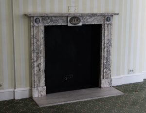 Front view of a marble fireplace mantel with a narrow shelf, carved jambs, and oval figural inlays at the center and corners.