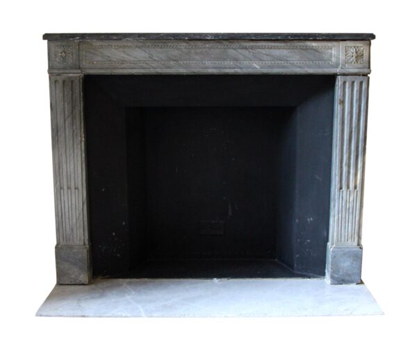Front view of a Louis XVI Regency marble mantel with fluted legs, floral corner motifs, and a dark shelf.