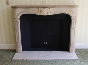 Louis XV carved mantel in tan limestone, shown frontally with a replacement hearth and an ornate central crest above the opening.