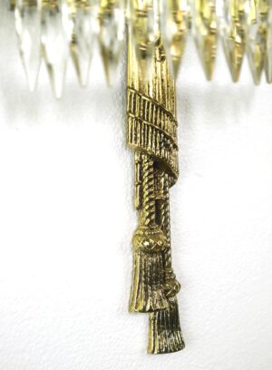 Close-up of a polished brass tassel detail on a crystal wall sconce, showing the ornate vertical ribbon and fringe motif.