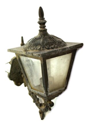 Victorian exterior wall lantern with a black cast aluminum frame and glass panels, shown at an angle with ornate top detailing and visible weathered wear.