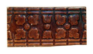 Brown ceramic decorative tile with raised geometric floral details and visible age-related chips and scrapes.