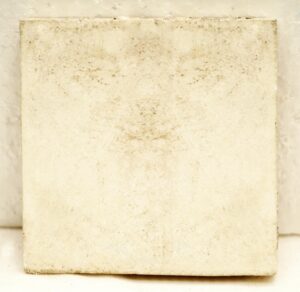 Square tan ceramic tile with a visibly worn surface and minor age-related imperfections.