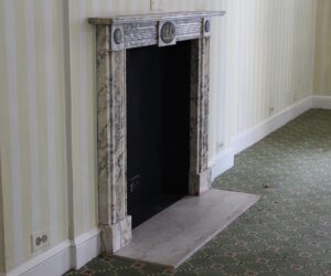 English Regency marble mantel with gray veining and figural cameo details, shown installed with a dark firebox opening and narrow shelf.
