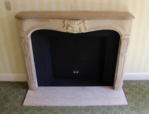 Louis XV carved limestone mantel with a replacement hearth and ornate central carving, shown installed with a tan top and dark firebox opening.
