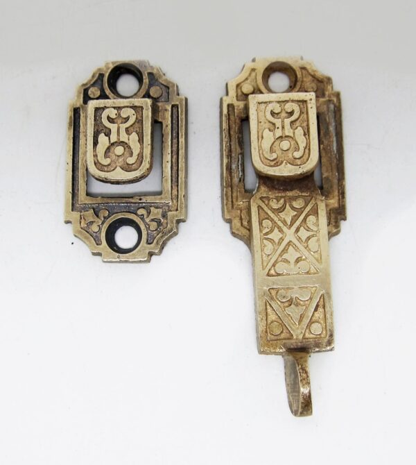 Ornate cast iron shutter latch with Victorian-style decorative panels and mounting holes, shown in two views with an attached hooked keeper and surface wear.