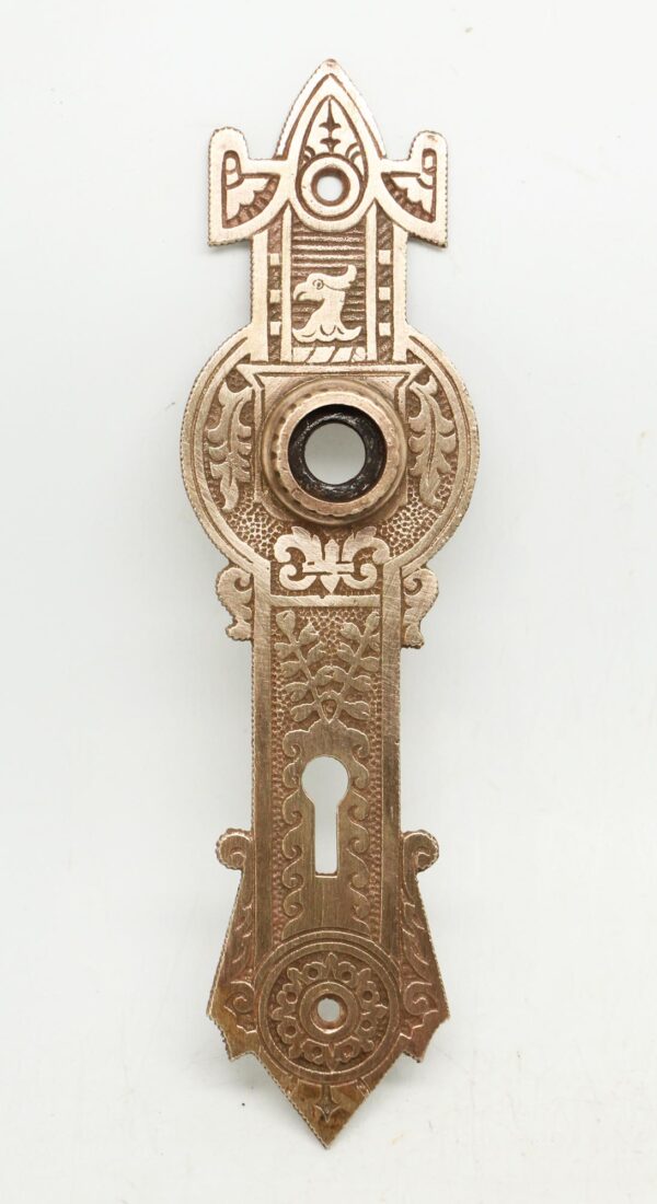 Ornate bronze door back plate with a keyhole opening and circular center hole, shown vertically with decorative cutouts and engraved detailing.
