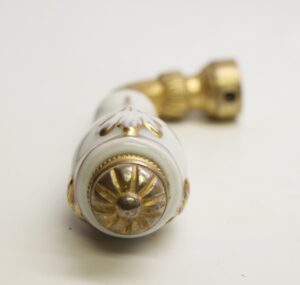 White porcelain faucet handle with ornate brass floral detailing, shown in close-up with the end cap facing forward and the mounting stem blurred behind it.