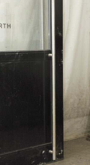 Black metal commercial door with one glass panel and a full-length pull bar, shown in a close-up of the latch side.