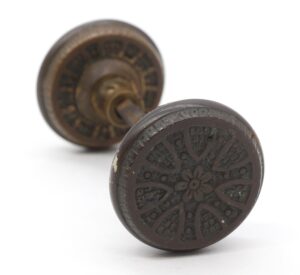 Pair of antique bronze passage door knobs, one front knob with a radial floral pattern and one blurred knob behind it.