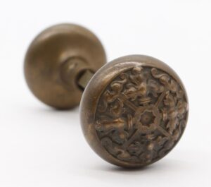Pair of antique bronze entry door knobs shown in close-up, with one knob featuring an ornate Flemish floral face and the other visible out of focus behind it.