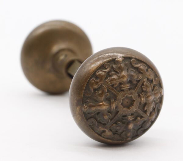 Pair of antique bronze entry door knobs shown in close-up, with one knob featuring an ornate Flemish floral face and the other visible out of focus behind it.