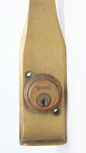 Close-up of an antique brass commercial door pull with a circular lock cylinder and two mounting holes visible.