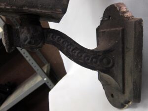 Close-up of an Arts & Crafts wrought iron wall sconce showing the curved arm and mounting plate with worn grayish green paint and a glass holder at the top.
