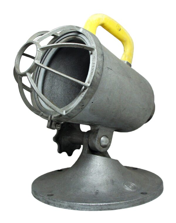 Gray steel spotlight with a protective front cage and yellow handle, shown in a side angle on a round base.