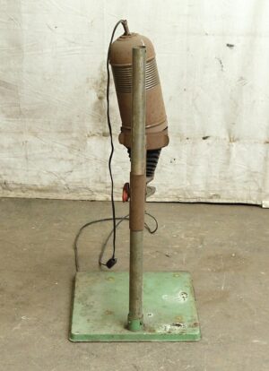 Vintage photo enlarger with a brown head on a green metal base, shown on a vertical stand with a cord hanging beside it and visible wear.