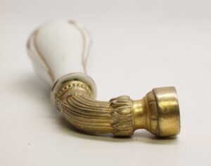 Ornate faucet handle with a white porcelain grip and brass stem shown in close-up from the side.