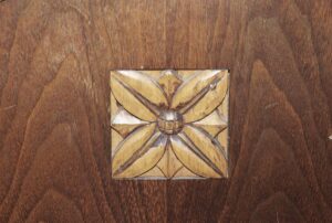 Close-up of a wooden chair back with a carved floral motif in dark and light wood tones.