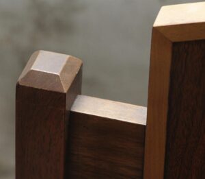 Close-up of a Mission style wooden chair back showing the carved top rail and dark and light wood tones.