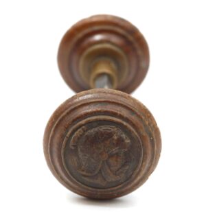 Pair of wooden passage door knobs with brass shanks, shown front on and slightly out of focus, one knob featuring a carved Greek head design and dark wood tone with age cracks.