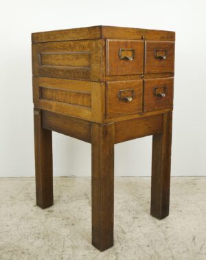 Four-drawer oak card catalog cabinet on four tall legs, with brass label pulls and visible metal tags on the front.