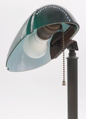 Close-up of a bankers desk lamp with a green exterior and white interior glass shade, a visible pull chain switch, and a fluted base.
