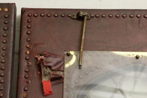 Close-up of a leather-studded door with a brass latch, showing a torn section and a clear protective panel over the stained glass area.