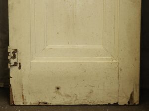 White wood door with one recessed panel, shown close up and weathered with a hinge on the left edge.
