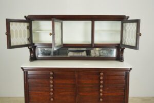 Antique mahogany dental cabinet with open embossed glass doors, a white milk glass top, a mirrored backsplash, and rows of drawers with round knobs.