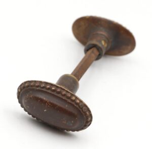 Pair of bronze oval door knobs connected by a spindle, with beaded edges and visible surface wear on the antique finish.