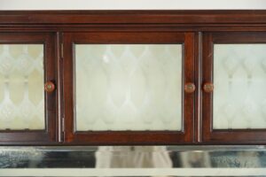 Upper section of a mahogany cabinet with three embossed glass doors, round bakelite-and-brass knobs, and a mirrored lower edge.