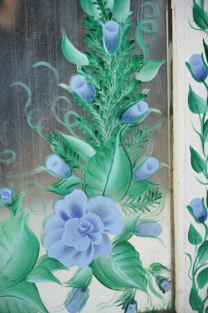 Close-up of a reclaimed window mirror with hand-painted blue roses and green leaves on the glass, showing part of the white original frame and the signed lower edge.