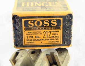 Close-up of a brass hinge package label reading Soss 212, with the hinge hardware partially visible below.