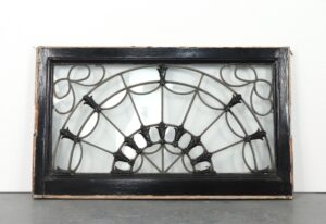 Antique fanlight transom window with a black-painted wood frame, radiating leaded glass design, and raised floral details, shown with mounting hardware and a visible crack in the glass.